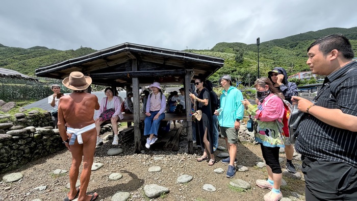 雅美族傳統「地下屋」由周克柱先生穿著傳統服飾親自介紹地下屋具有氣候適應與防風智慧：主屋（Vahay）向下深掘、半掩於地穴中，僅露出屋頂斜面，能有效抵禦蘭嶼強烈颱風與冬季東北季風，並具冬暖夏涼的調溫效果。圖片
